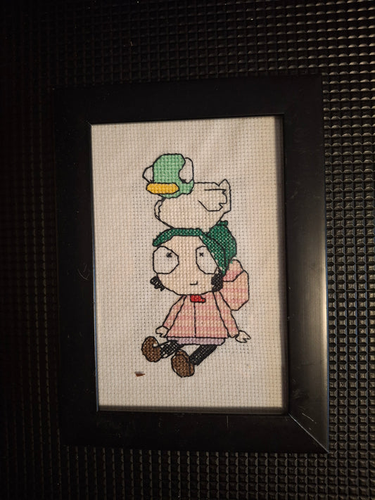 Girl with a duck sat her head  cross stitched design in frame